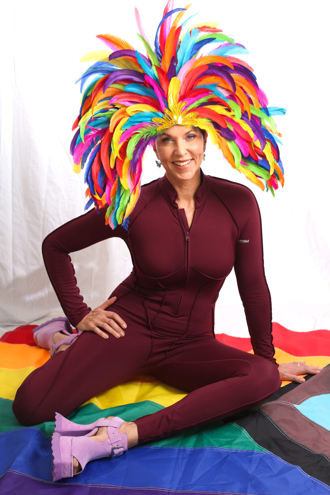 Grace Walcott seated on a rainbow flag wearing a large multicolored feather headdress
