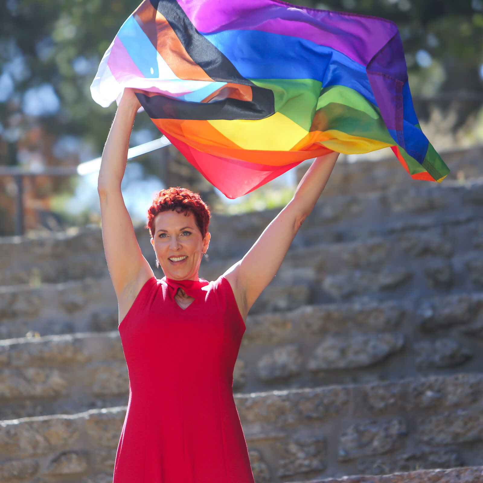 Grace Walcott raising a Progress Pride flag overhead, smiling toward camera