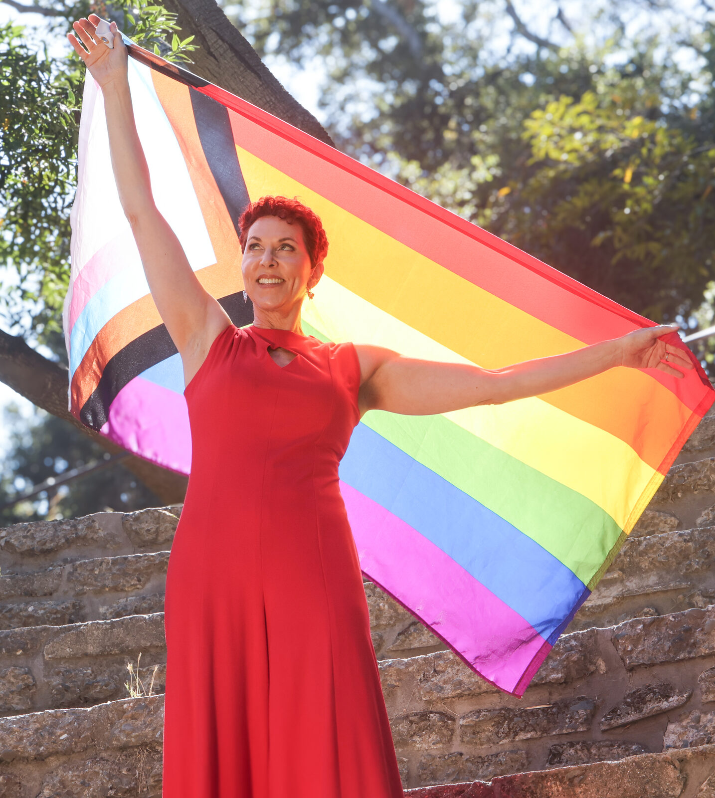 Grace Walcott in a red gown holding a Progress Pride flag wide above her, on stone amphitheater steps