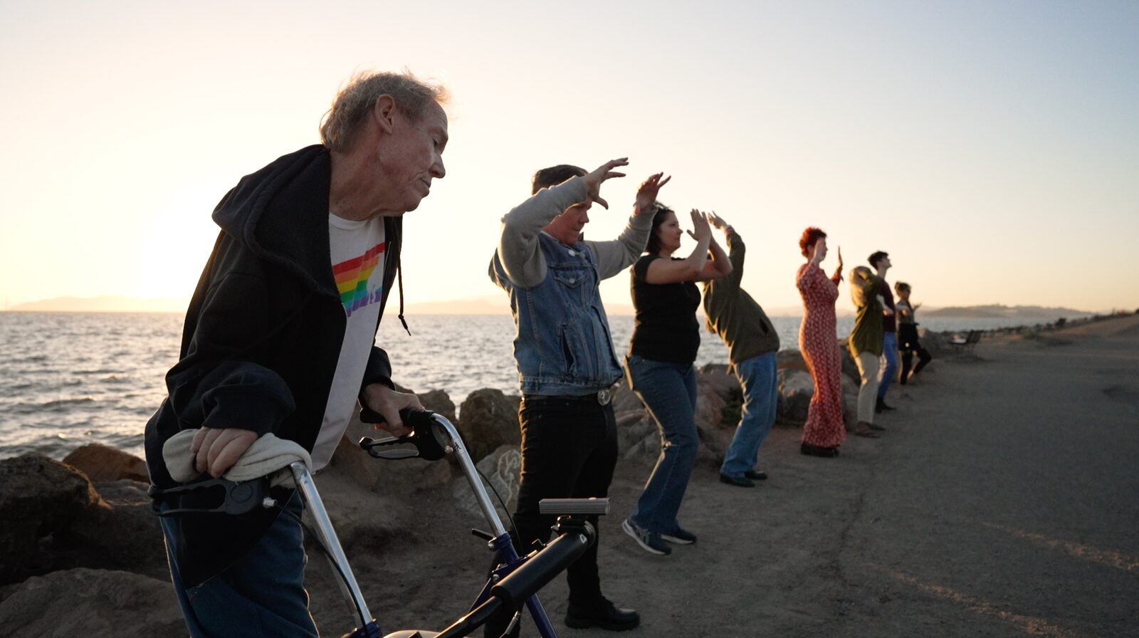 Participants including an elder with a walker gather on the shoreline